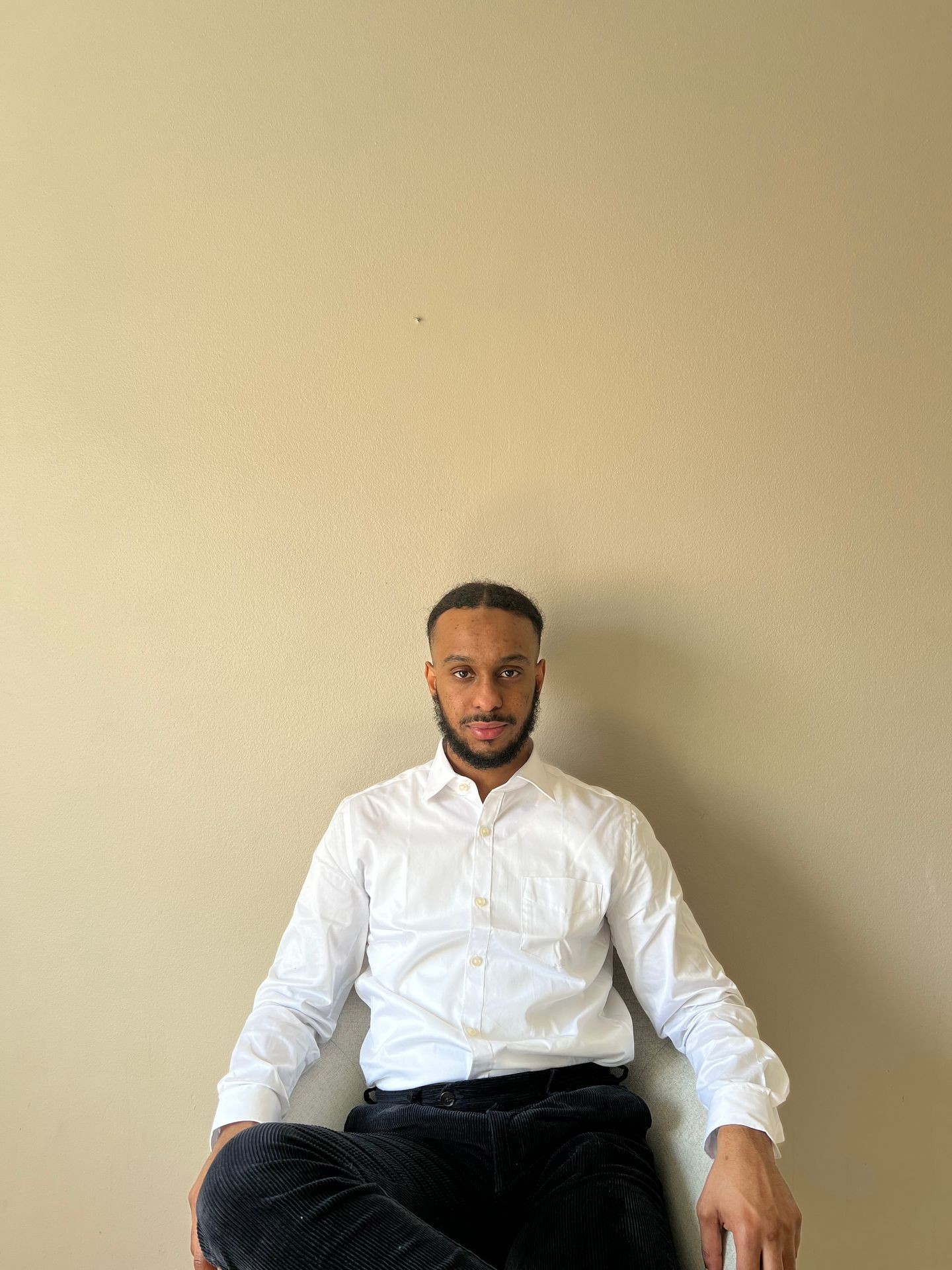 Person sitting against a beige wall wearing a white dress shirt and dark pants.